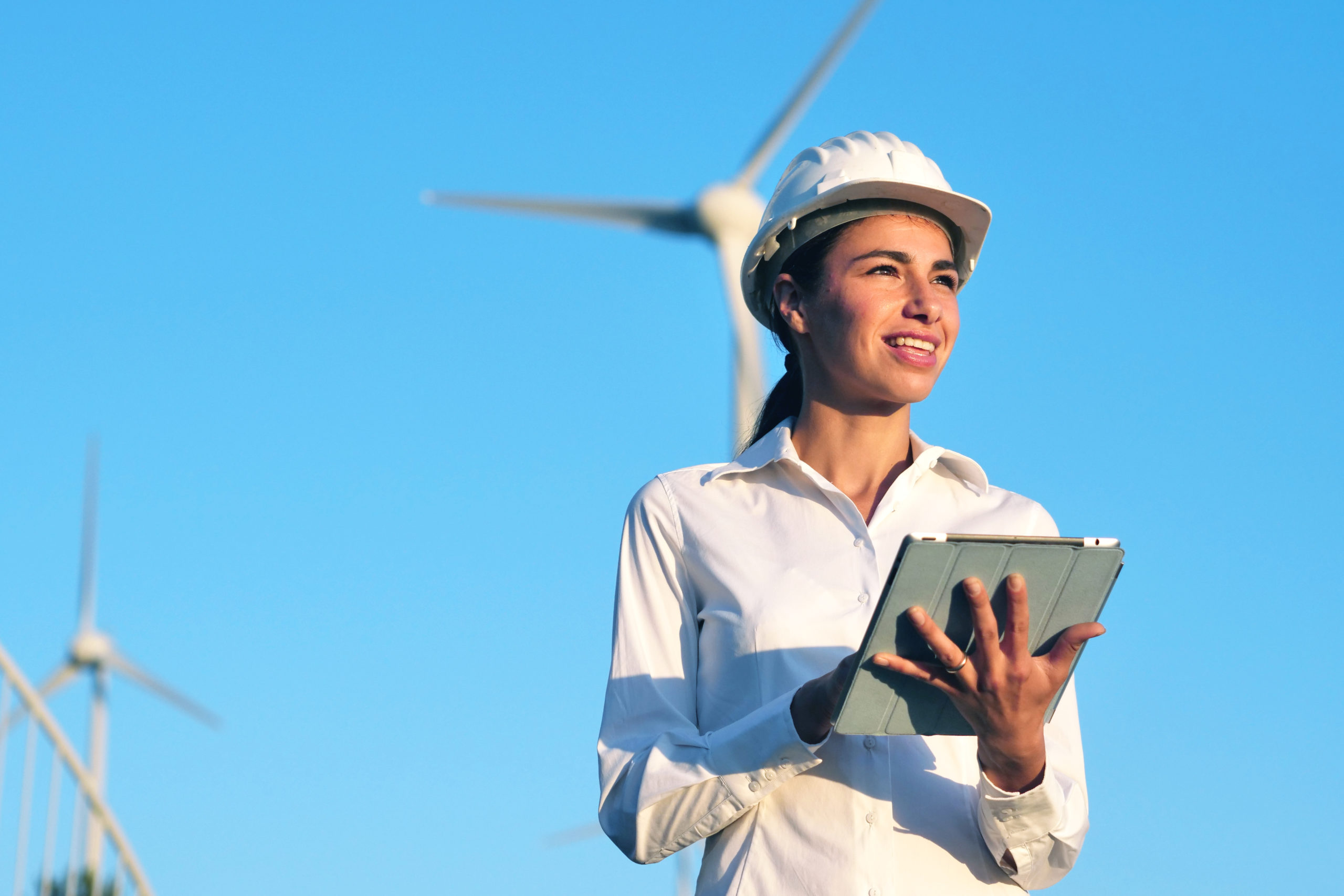 Featured Image. A female engineer in a white hard hat with a notepad with windturbines in the background