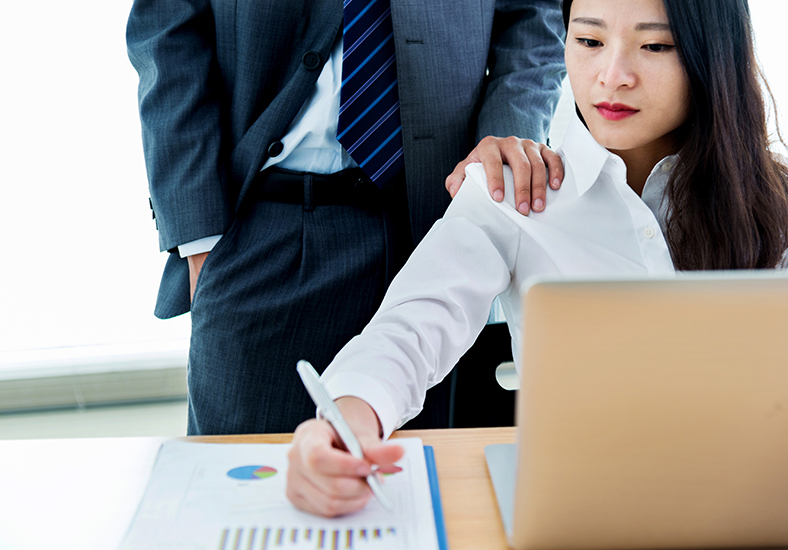 Featured Image. Image of female office worker working on laptop with intimidating male co-worker presence